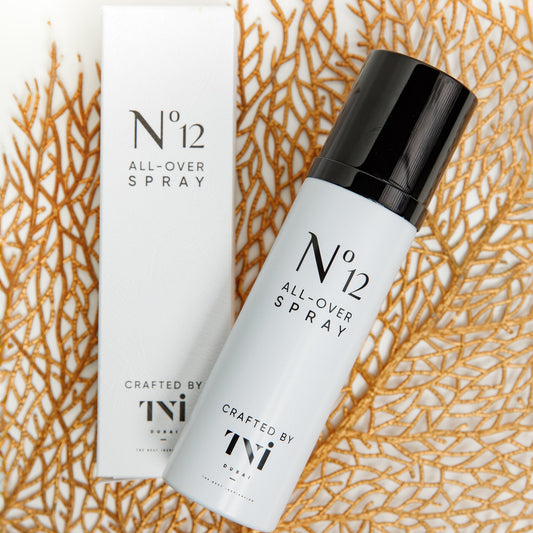 A white spray bottle labeled 'N°12 ALL OVER SPRAY' positioned next to its packaging box, both featuring minimalistic design, placed against a textured orange backdrop.